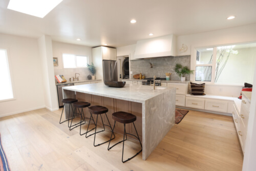 Mid-century modern cream shaker kitchen cabinet design with a large custom kitchen hood and medium wood-toned island cabinets with beautiful gray stone waterfall countertops