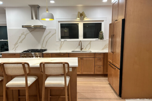 L-shaped mid-tone brown shaker kitchen with paneled refrigerator, large island with quartz countertops and matching slab backsplash