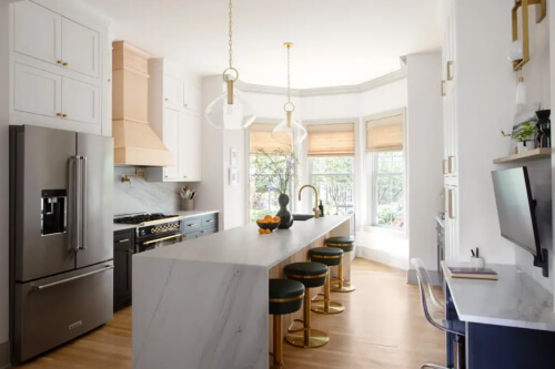 Stacked white inset shaker cabinets with black island cabinets, creamy waterfall countertops, funky glass and gold light fixtures, a natural wood kitchen hood