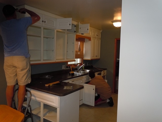 two men removing cabinet doors and disconnecting sink plumbing in kitchen with white cabinets and black countertops