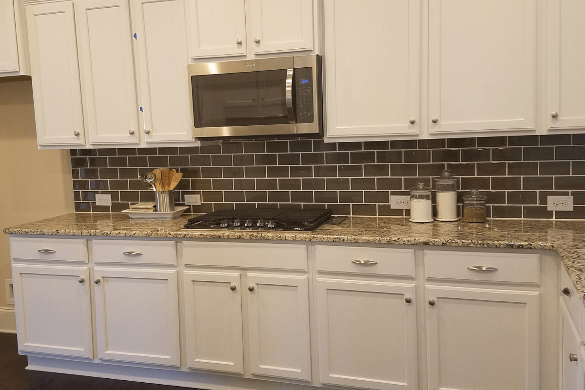 Cooktop wall of a white shaker kitchen design with partial overlay kitchen cabinets, granite countertops and a stainless steel microwave.