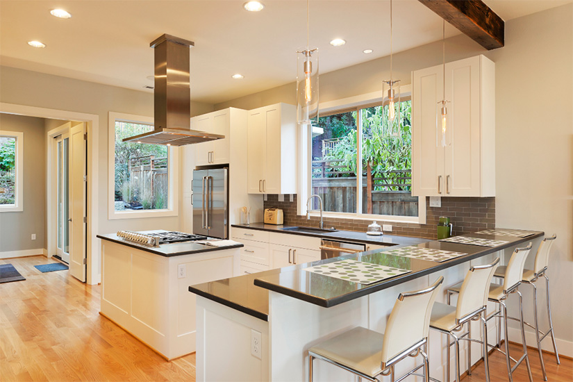 White shaker L-shaped industrial kitchen with gray quartz countertops, a wood beam and stainless-steel island hood.